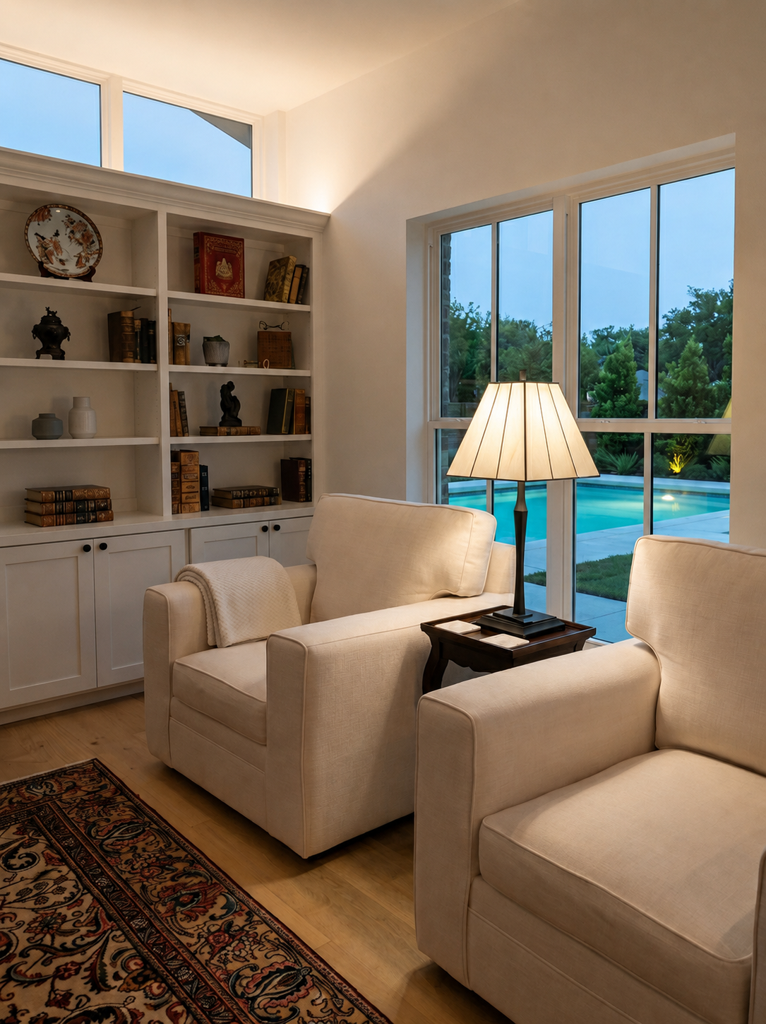 Hollywood Park sitting room with built-in bookshelves, linen armchairs and pool view at dusk